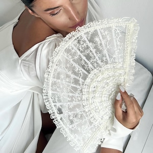 May include: A white lace fan with a scalloped edge. The fan is made of delicate fabric and has a delicate floral pattern.