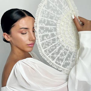 May include: A woman in a white dress holds a white lace fan in front of her face. The fan is decorated with delicate embroidery and has a scalloped edge.