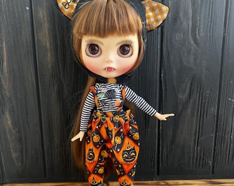 May include: A doll with brown hair wearing a black and orange Halloween costume with a black cat hat that says "Boo!" on it. The doll is wearing overalls with a black cat and pumpkin pattern.