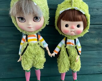 May include: Two dolls wearing green fuzzy outfits with striped shirts and green shoes. The dolls have different hair colours and facial expressions.