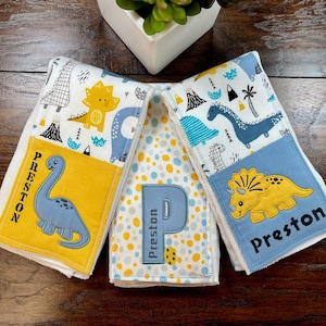 May include: Three white burp cloths with blue and yellow dinosaur designs. Each cloth has a yellow pocket with a dinosaur and the name "Preston" embroidered on it.