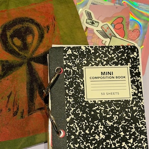 Può includere: Un mini quaderno di composizione con copertina maculata bianca e nera, tenuto insieme da due anelli metallici argentati. Accanto si trova una sacca con coulisse verde oliva con una figura aliena nera su sfondo rosso e una bustina olografica di adesivi.