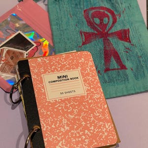 Può includere: Un mini quaderno con copertina maculata rosa e bianca, dorso nero e due anelli metallici. Accanto si trova un sacchetto turchese con coulisse decorato con un simbolo ankh alieno rosso e una bustina olografica rosa contenente vari adesivi.