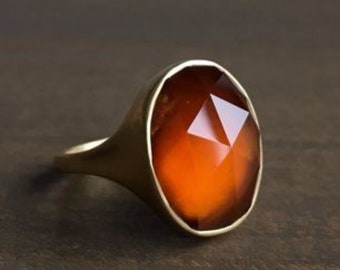 Puede incluir: Un anillo de oro con una piedra preciosa naranja ovalada y facetada. El anillo tiene un diseño simple y elegante con una banda lisa y pulida. La piedra preciosa es el punto focal, captando la luz y mostrando un color cálido.