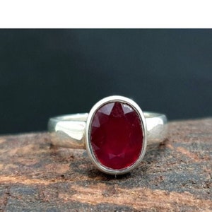 May include: A silver ring featuring a large, oval-cut ruby gemstone. The ruby is a deep, rich red color and is set in a simple, polished silver band. The ring is displayed on a textured wooden surface.