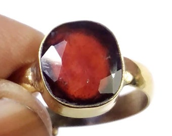 Puede incluir: Un anillo de oro con una gran gema roja oscura, tallada en forma ovalada y facetada. La banda y el engaste del anillo son dorados, creando un contraste con la gema roja. El anillo se muestra sobre un fondo blanco.