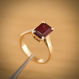 Op de afbeelding: Een gouden ring met een rechthoekige, dieprode edelsteen. De ring heeft een gladde, gepolijste band en de edelsteen wordt vastgehouden door vier klauwen. De ring wordt door een pincet tegen een vage achtergrond gehouden.