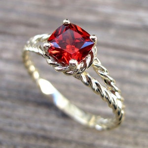 Könnte beinhalten: Ein goldfarbener Ring mit einem quadratischen, leuchtend roten Edelstein. Das Band weist ein gedrehtes Seilmuster auf, und der Edelstein wird von vier kleinen Krappen gehalten. Der Ring ist vor einem verschwommenen Holzgrund platziert.