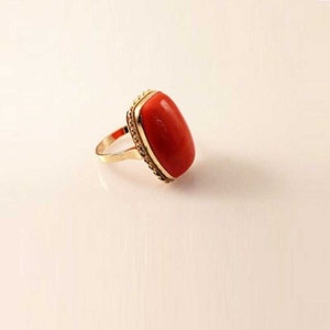 May include: A gold ring featuring a large, oval-shaped, red gemstone. The gemstone is set in a gold bezel with a decorative edge. The ring band is also gold, creating a cohesive and elegant design. The ring is displayed on a white surface.