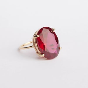 Könnte beinhalten: Ein goldener Ring mit einem großen, oval geschliffenen, rubinroten Edelstein. Der Ring hat einen einfachen Band und eine Vier-Zacken-Fassung, die den leuchtenden Stein sichert. Die Facetten des Edelsteins fangen das Licht ein.