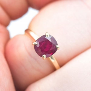 Puede incluir: Un anillo de oro con una gran gema de rubí tallada en cojín. El rubí es de un color rojo intenso y está sujeto por cuatro garras de oro. La banda del anillo es de oro pulido simple.