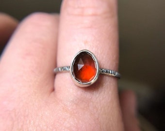 Könnte beinhalten: Ein Silberring mit einem facettierten, tropfenförmigen, rot-orangen Edelstein. Das Ringband hat ein strukturiertes Design. Der Edelstein ist in einer silbernen Lünette gefasst.