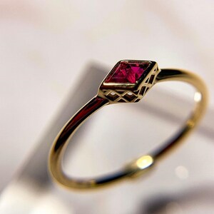 May include: A gold ring featuring a vibrant, square-cut ruby gemstone. The ruby is set within a geometric, openwork bezel. The ring's band is a slender, polished gold. The design is elegant and minimalist, suitable for everyday wear or special occasions.