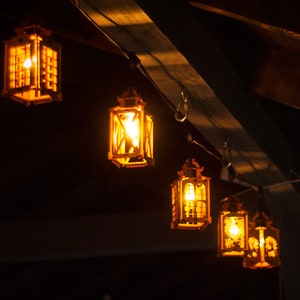 以下が含まれることがあります： 木製の天井から吊り下げられた、暖かく光る光を放つ5つの木製提灯の連なり。提灯は薄い茶色の木で作られており、シンプルで素朴なデザインです。