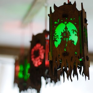 May include: A set of three wooden Halloween lanterns with intricate cutouts. The lanterns are illuminated from within, casting a warm glow. The lantern in the foreground features a green light and a cutout of a haunted house.