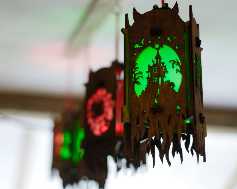 May include: A set of three wooden Halloween lanterns with intricate cutouts. The lanterns are illuminated from within, casting a warm glow. The lantern in the foreground features a green light and a cutout of a haunted house.