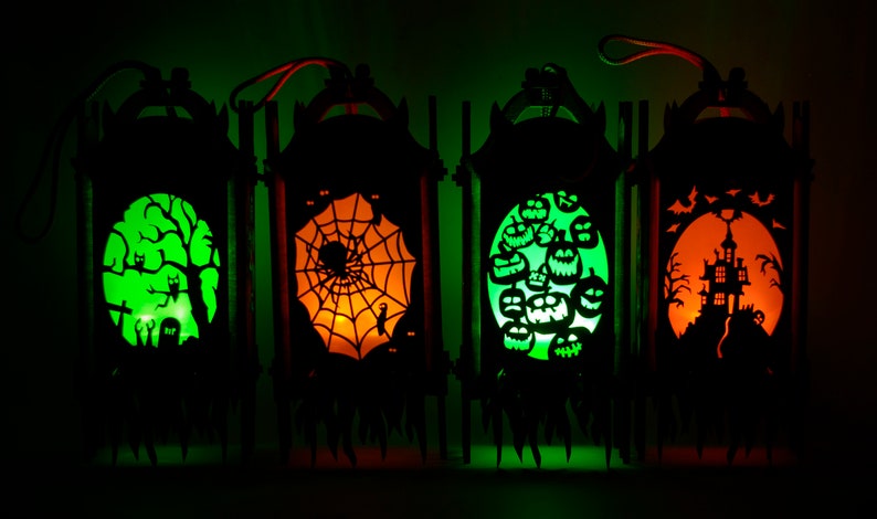 May include: Four black wooden Halloween lanterns with different designs. The lanterns are lit from within, casting a warm glow. The designs include a spooky tree with owls, a spider web, a group of jack-o'-lanterns, and a haunted house.