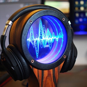 May include: A wooden headphone stand with a circular display showing a blue sound wave. The stand has a dark frame and copper-colored rivets. Black headphones with silver accents rest on the stand.