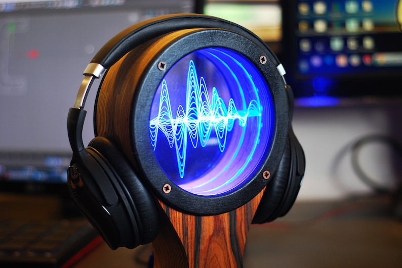 May include: A wooden headphone stand with a circular display showing a blue sound wave. The stand has a dark frame and copper-colored rivets. Black headphones with silver accents rest on the stand.