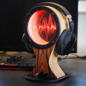 May include: A wooden headphone stand with a circular design featuring a red illuminated sound wave pattern. The stand has a black frame and a wooden base. Black headphones rest on the stand.