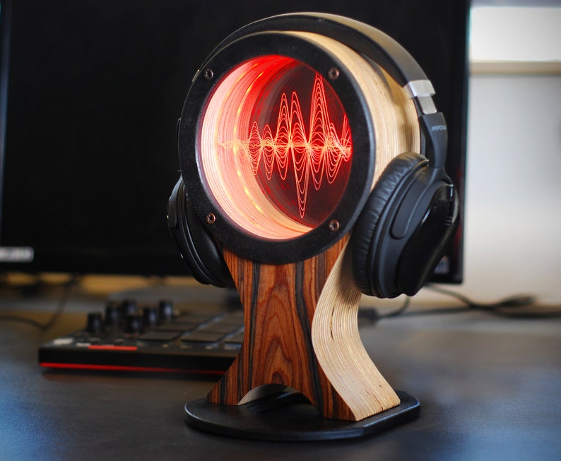 May include: A wooden headphone stand with a circular design featuring a red illuminated sound wave pattern. The stand has a black frame and a wooden base. Black headphones rest on the stand.