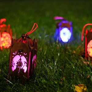 May include: Four wooden Halloween lanterns with different coloured lights. The lanterns are in the shape of houses and have spooky designs cut out of them. The lanterns are sitting on a bed of green grass.