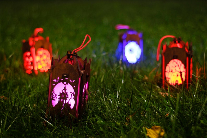 May include: Four wooden Halloween lanterns with different coloured lights. The lanterns are in the shape of houses and have spooky designs cut out of them. The lanterns are sitting on a bed of green grass.
