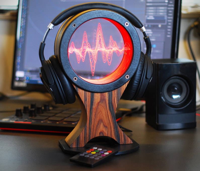 May include: A wooden headphone stand with a circular display showing a red sound wave. Black headphones rest on the stand. A remote control with colorful buttons sits in front. The stand has a unique wood grain pattern.