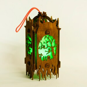 May include: A wooden lantern with a green glow, featuring a spooky tree and graveyard scene. The lantern has a red string for hanging.