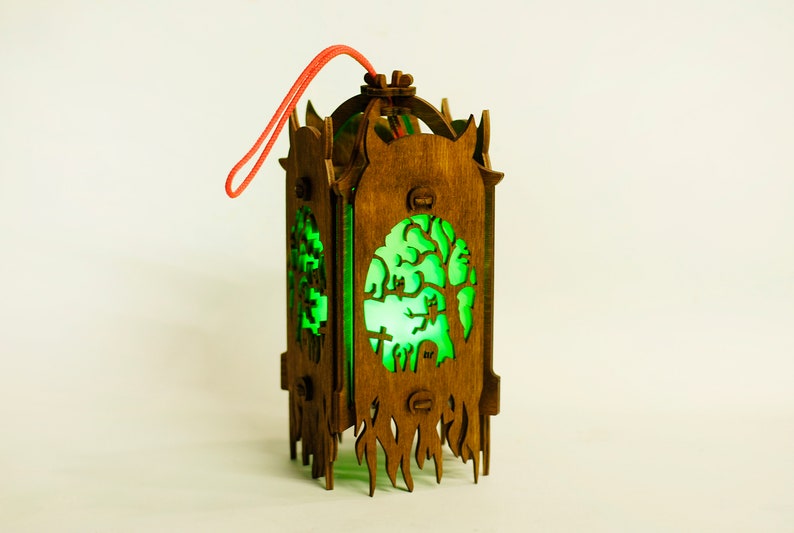 May include: A wooden lantern with a green glow, featuring a spooky tree and graveyard scene. The lantern has a red string for hanging.