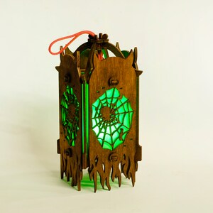 May include: A wooden lantern with a green glass panel featuring a spiderweb design. The lantern has a red string handle and a decorative, flame-like base.
