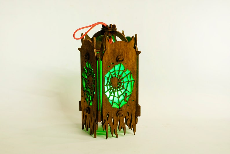 May include: A wooden lantern with a green glass panel featuring a spiderweb design. The lantern has a red string handle and a decorative, flame-like base.
