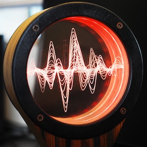 May include: A unique, circular audio visualizer with a wooden frame and a black rim. The display shows a glowing red waveform pattern. The design is mounted on a wooden base, creating a modern, artistic piece.