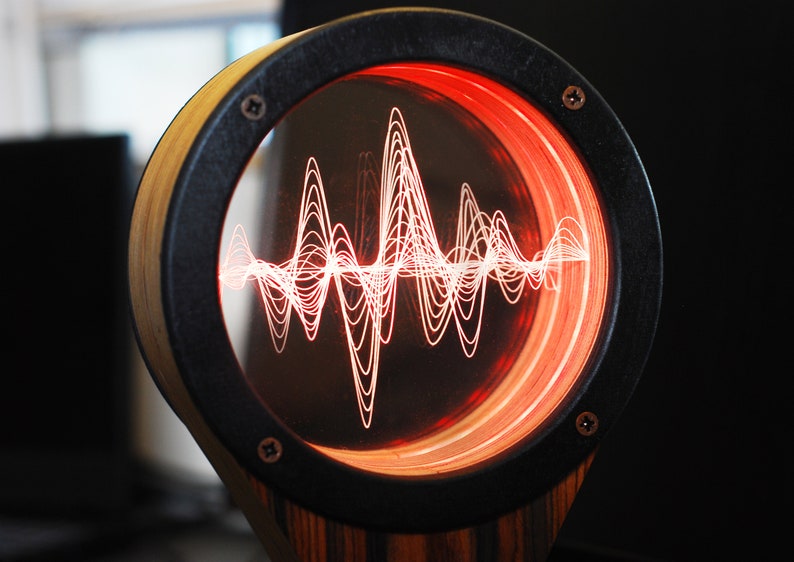 May include: A unique, circular audio visualizer with a wooden frame and a black rim. The display shows a glowing red waveform pattern. The design is mounted on a wooden base, creating a modern, artistic piece.