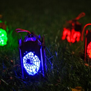 May include: Four Halloween lanterns with different designs and colours. The lanterns are lit up with green, red, and blue lights. The lanterns are sitting on a bed of green grass.