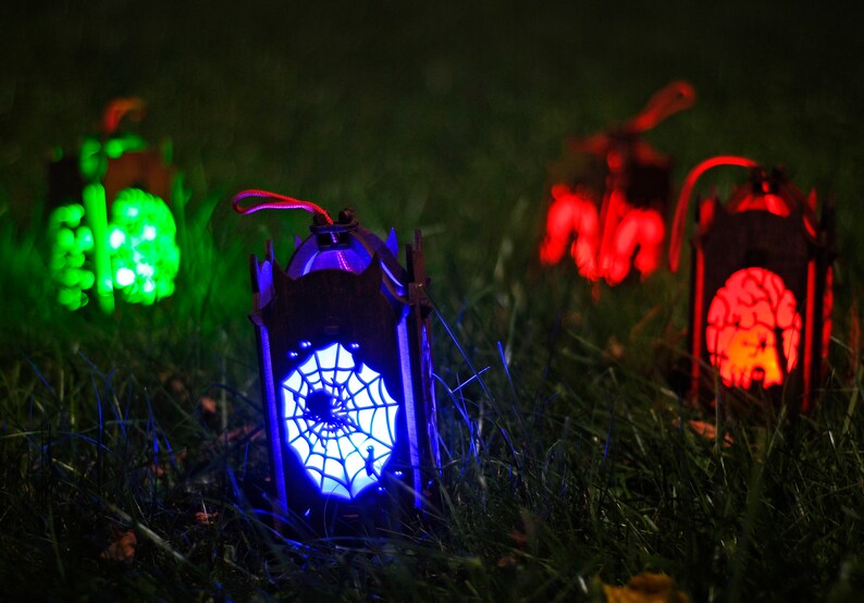 May include: Four Halloween lanterns with different designs and colours. The lanterns are lit up with green, red, and blue lights. The lanterns are sitting on a bed of green grass.
