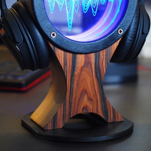 May include: Headphone stand with a wooden base and a circular display showing a blue sound wave. The stand has a dark wood grain pattern and holds a pair of black headphones. The display is framed in black.