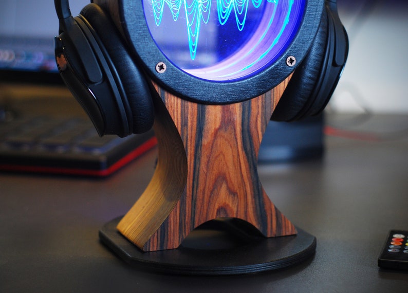 May include: Headphone stand with a wooden base and a circular display showing a blue sound wave. The stand has a dark wood grain pattern and holds a pair of black headphones. The display is framed in black.