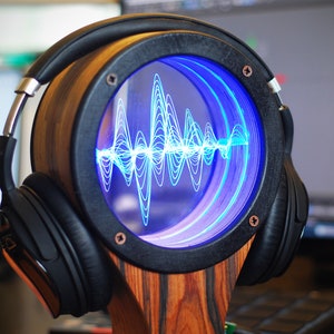 May include: A wooden headphone stand with a circular display showing a blue sound wave pattern. The stand has a black frame and is holding a pair of black headphones. The stand is made of wood with a zebra-like pattern.