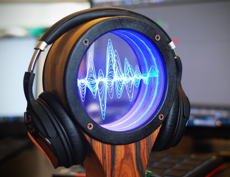 May include: A wooden headphone stand with a circular display showing a blue sound wave pattern. The stand has a black frame and is holding a pair of black headphones. The stand is made of wood with a zebra-like pattern.
