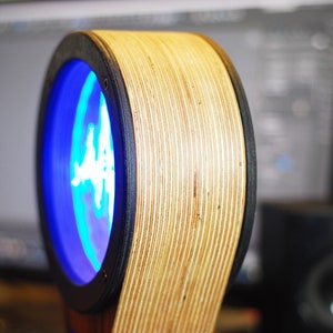 May include: A unique, circular wooden speaker with a blue illuminated center. The speaker has a layered wood design with a black trim. The speaker is designed to be a modern and stylish addition to any home audio setup.