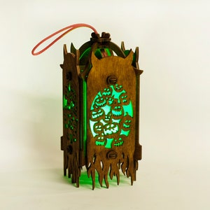 May include: A wooden lantern with a green glow and a carved design of jack-o'-lanterns. The lantern has a red string attached to the top for hanging.