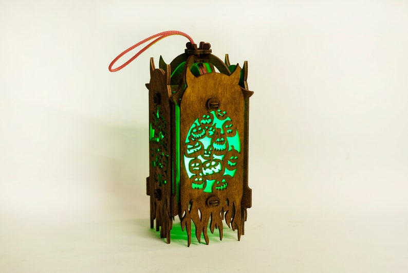 May include: A wooden lantern with a green glow and a carved design of jack-o'-lanterns. The lantern has a red string attached to the top for hanging.