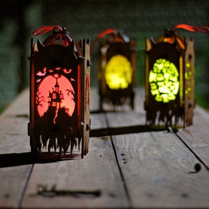 May include: Three wooden Halloween lanterns with intricate cutouts. The lanterns are lit from within, casting a warm glow. The lantern on the left has a red glow and depicts a haunted house, while the other two lanterns have a yellow glow and depict different designs.