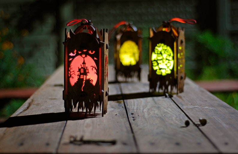 May include: Three wooden Halloween lanterns with intricate cutouts. The lanterns are lit from within, casting a warm glow. The lantern on the left has a red glow and depicts a haunted house, while the other two lanterns have a yellow glow and depict different designs.