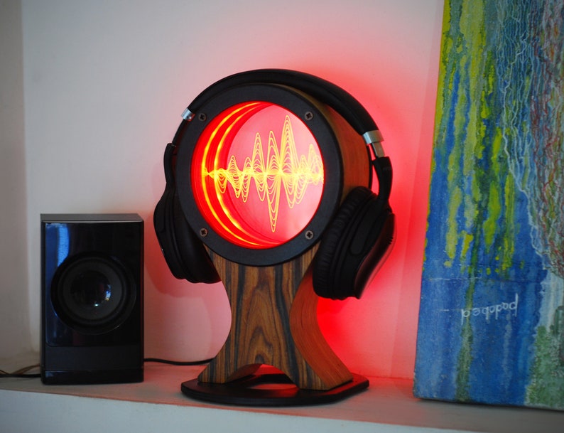 May include: A unique audio visualizer with a wooden base and a circular frame. The frame holds a pair of black headphones and a red-lit display showing a yellow sound wave pattern. A black speaker sits to the left.