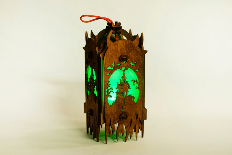 May include: A wooden lantern with a green glow, featuring a silhouette of a haunted house and bats. The lantern has a red string for hanging.