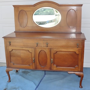 May include: A vintage wooden sideboard with a beveled mirror backsplash. The sideboard has three doors with brass hardware and two drawers with brass knobs. The sideboard is finished in a warm brown color and has cabriole legs.