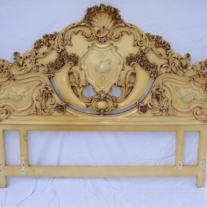 May include: Cream-colored, ornate headboard with intricate carvings and floral accents. The headboard features a shell-shaped top and a shield-like center design with painted flowers. The frame is supported by two vertical posts.