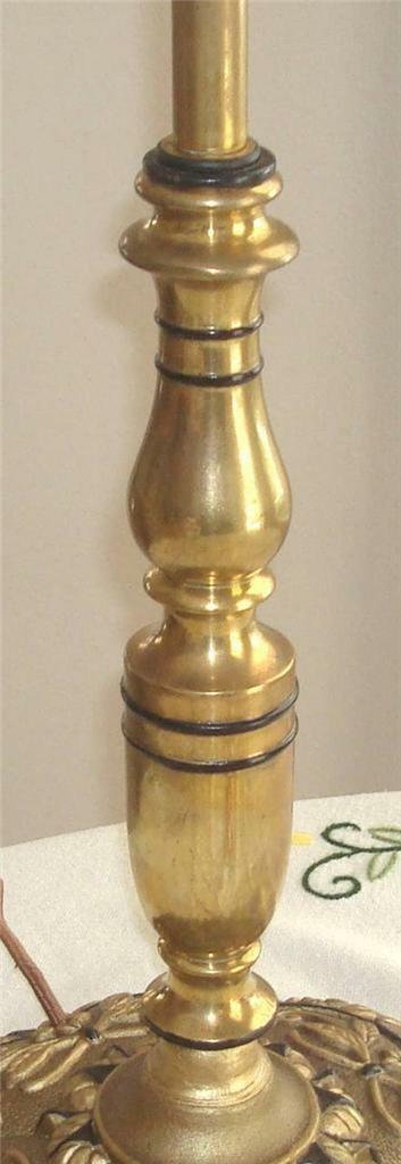 May include: A gold-toned lamp base with black accents. The base is a tall, slender cylinder with a bulbous middle section. The base has a decorative, ornate design at the bottom.